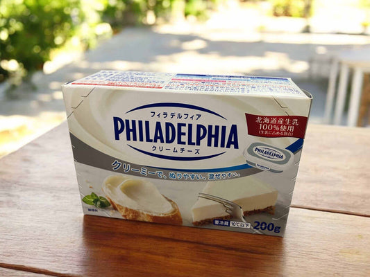 Philadelphia cream cheese 200g box with Japanese text, made from 100% Hokkaido milk, displayed on wooden table outdoors