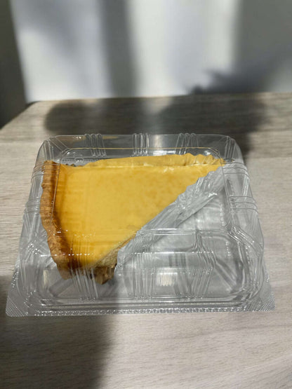 Slice of triple cheese tart in clear plastic container on wooden surface with natural light shadows